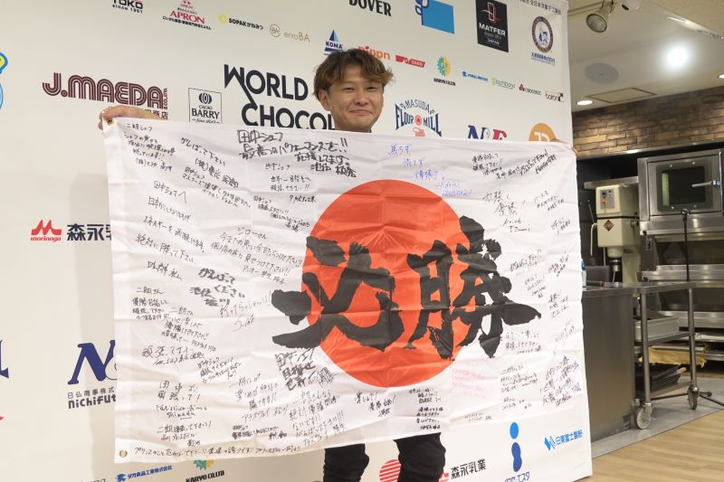 「ワールド チョコレート マスターズ2022」日本代表壮行会での田中二朗シェフ。応援旗の寄せ書きには師匠や歴代選手、仲間達のメッセージが